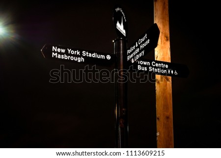 Signpost to Rotherham Town Centre, Bus Station, Library, Police Station, Court, Riverside House & Rotherham United Stadium (New York) - at night, with a street light background