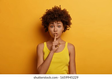 Silence And Secret Concept. Surprised African American Woman Presses Index Finger To Lips, Asks Be Quiet, Tells Very Private Information, Wears Casual Yellow Vest, Poses In Studio, Says Shush Or Hush