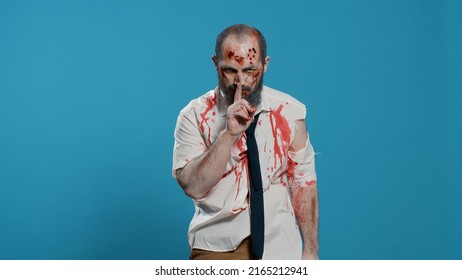 Silent creepy zombie making shush secrecy hand gesture on blue background. Dangerous walking dead corpse with deep and bloody wounds being confidential while smirking at camera.