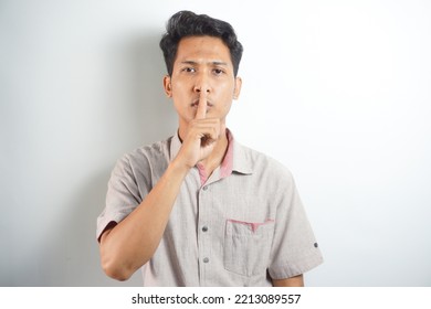 Silent Hand Symbol Or Shhh (ssst). Portrait Of Asian Man Glaring While Looking At Camera With Index Finger In Front Of Mouth. Indonesian Man Wearing A Hoodie On A White Background