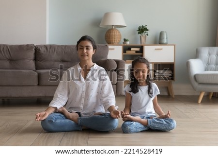 Silent Indian woman and preschooler 5s serene daughter meditating seated in lotus position on warm floor in modern living room. Good life habit, healthy lifestyle, yoga practice with children at home