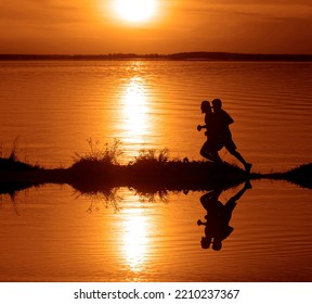 Silhouette Of 2 Two Men Running Together On A Sunset On Lake Coast. Side View Of 2 Men Silhouette Running On The Sea Shore. Couple Running On The Beach At Sunrise.