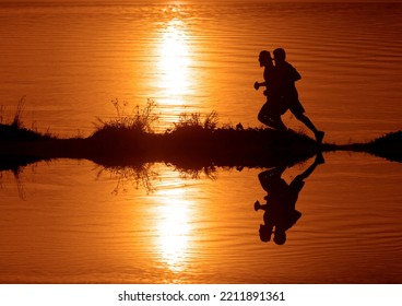 Silhouette Of 2 Two Men Running Together On A Sunset On Lake Coast. Side View Of 2 Men Silhouette Running On The Sea Shore. Couple Running On The Beach At Sunrise.