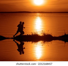 Silhouette Of 2 Two Men Running Together On A Sunset On Lake Coast. Side View Of 2 Men Silhouette Running On The Sea Shore. Couple Running On The Beach At Sunrise.