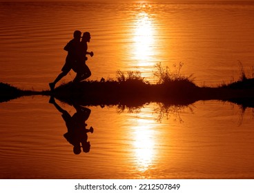 Silhouette Of 2 Two Men Running Together On A Sunset On Lake Coast. Side View Of 2 Men Silhouette Running On The Sea Shore. Couple Running On The Beach At Sunrise.