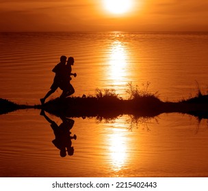 Silhouette Of 2 Two Men Running Together On A Sunset On Lake Coast. Side View Of 2 Men Silhouette Running On The Sea Shore. Couple Running On The Beach At Sunrise.