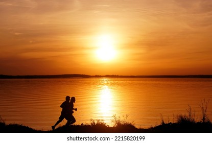 Silhouette Of 2 Two Men Running Together On A Sunset On Lake Coast. Side View Of 2 Men Silhouette Running On The Sea Shore. Couple Running On The Beach At Sunrise.