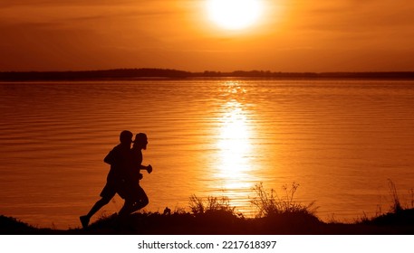 Silhouette Of 2 Two Men Running Together On A Sunset On Lake Coast. Side View Of 2 Men Silhouette Running On The Sea Shore. Couple Running On The Beach At Sunrise.