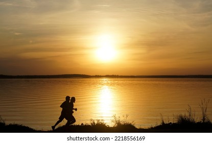 Silhouette Of 2 Two Men Running Together On A Sunset On Lake Coast. Side View Of 2 Men Silhouette Running On The Sea Shore. Couple Running On The Beach At Sunrise.