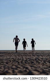 Silhouette of 3 boys of varying sizes running into the distance on beach at sunset beneath blue sky in summer