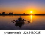 Silhouette of a 60-year-old Caucasian man with a fishing rod and a poodle dog in a wooden rowing boat with oars in his hands against the background of a sunset.