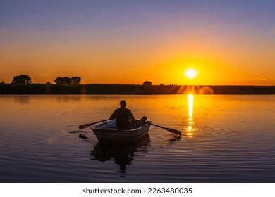 Silhouette of a 60-year-old Caucasian man with a fishing rod and a poodle dog in a wooden rowing boat with oars in his hands against the background of a sunset.