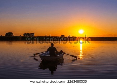Silhouette of a 60-year-old Caucasian man with a fishing rod and a poodle dog in a wooden rowing boat with oars in his hands against the background of a sunset.