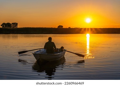 Silhouette de un hombre caucásico de 60 años con un perro caniche en un tradicional bote remo con remos en las manos en el fondo de una puesta de sol.