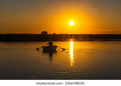 The silhouette of a 60-year-old fisherman in a wooden rowing boat with oars against the background of sunset at dusk. The yellow sun is reflected by the path on the water - Powered by Shutterstock