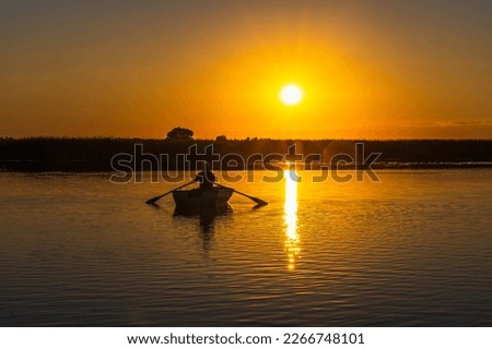 The silhouette of a 60-year-old fisherman in a wooden rowing boat with oars against the background of sunset at dusk. The yellow sun is reflected by the path on the water