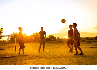 Silhouette Action Sport Outdoors Of A Group Of Kids Having Fun Playing Soccer Football For Exercise In Community Rural Area Under The Twilight Sunset. Poor And Poverty Children In Development Country.