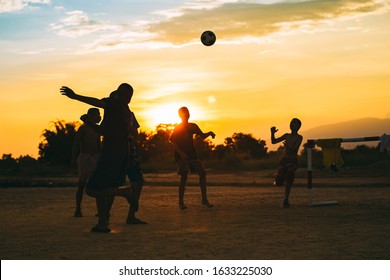 Silhouette Action Sport Outdoors Of A Group Of Kids Having Fun Playing Soccer Football For Exercise In Community Rural Area Under The Twilight Sunset. Poor And Poverty Children In Development Country.