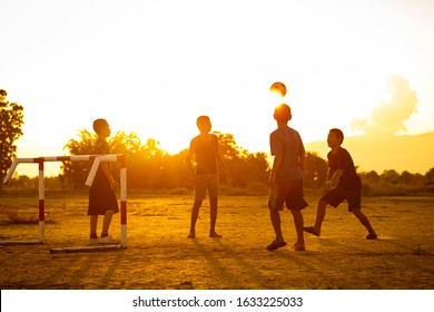 Silhouette Action Sport Outdoors Of A Group Of Kids Having Fun Playing Soccer Football For Exercise In Community Rural Area Under The Twilight Sunset. Poor And Poverty Children In Development Country.