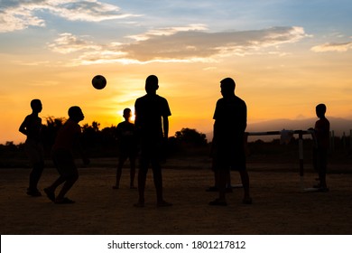 Silhouette Action Sport Outdoors Of A Group Of Kids Having Fun Playing Soccer Football For Exercise In Community Rural Area Under The Twilight Sunset. Poor And Poverty Children In Development Country.