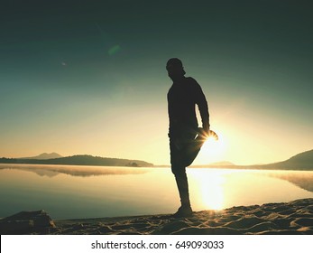 Silhouette of active man exercising  and stretching on the lake beach at sunrise. Healthy lifestyle. Alone young fitness man exercise  at morning beach