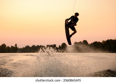silhouette of active man flying over splashing wave on wakeboard holding on to the rope. Wakeboarding and water sports activity.