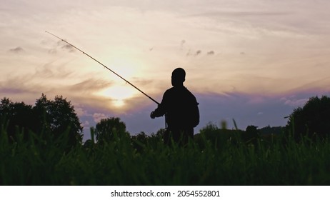 Silhouette of Adult Male Trying to Catch a Fish using Spinning Technique with Carbon Fiber Fishing Rod in Rays of Sun during Sunset