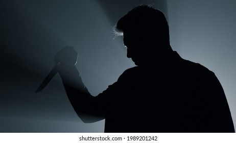 Silhouette of aggressive man standing with knife in hand indoors. Dangerous criminal using weapon in floodlight background. Scary gangster attacking with dagger in dark space. Crime concept.