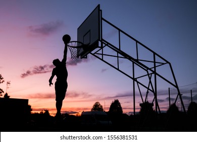 silhouette alone dunking man at sunset. basketball player scoring. Young silhouetted man jumping high to the target basketball hoop achieves to score with a flying slam dunk at sunset.