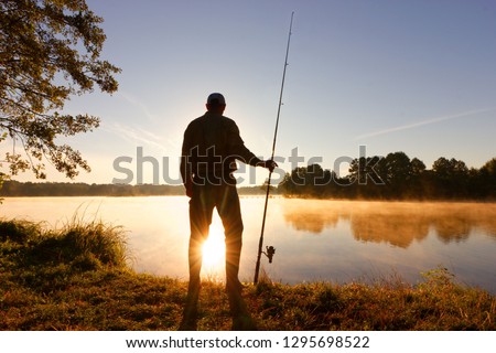 Silhouette of angler standing on the lake shore during misty sunrise
