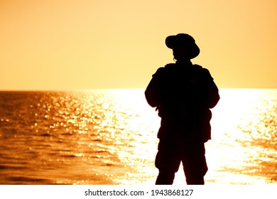 Silhouette Of Army Infantry Soldier, Coast Guard Fighter In Boonie Hat, Standing On Shore, Sunset And Ocean Horizon On Background. Border Guard Service Soldier Patrolling Coastline, Standing On Beach