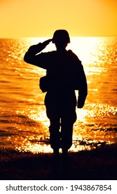 Silhouette of army soldier, Marines fighter in helmet and ammunition saluting while standing on seashore at sunset time. Coast guard fighter, special operations forces soldier honoring fallen comrades