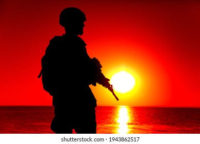 Silhouette Of Army Special Forces Rifleman, Marines, Coast Guard Or Anti-terrorist Squad Soldier In Ammunition And Helmet, Armed Service Rifle, Standing On Ocean Shore, Patrolling Seacoast At Sunset