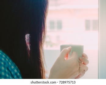 Silhouette Of Asian Girl In Blue Flannel Shirt Hold Hot Coffee For Drink In Morning Time And Look To Outside With Soft Focus Foreground And Background