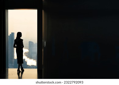 Silhouette Asian woman standing by the window at skyscraper building in metropolis and looking cityscape at summer sunset. Beautiful girl enjoy urban lifestyle activity in the city on holiday vacation
