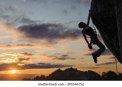 Silhouette Of Athletic Woman Rock Climber Climbing Steep Rock Wall With Rope Against Amazing Sunset Sky In The Mountains. Girl Is Hanging On One Hand And Holding Hand In Magnesium Bag.