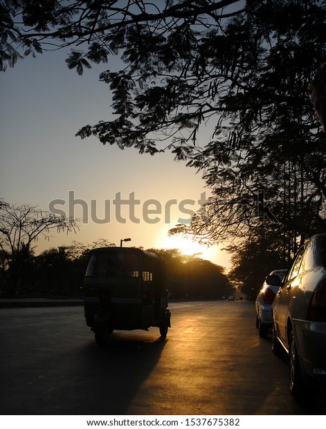 Silhouette of an auto rickshaw on a road in Dhaka,\
Bangladesh. The rickshaw has the setting sun behind. The golden\
sunlight reflects off the road. Local tuk tuk transport in Dhaka\
Bangladesh at sunset