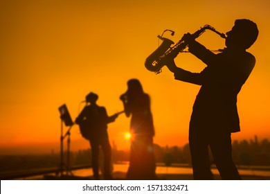 Silhouette autumn or summer scene of saxophone musician man showing with blurry jazz trio band and twilight or sunset city scape background. Image for happy new year party or celebration concept.