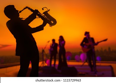 Silhouette Autumn Or Winter Scene Of Saxophone Musician Man Showing With Blurry Jazz Trio Band And Twilight Or Sunset Cityscape Background. Image For Happy New Year Party Or Celebration Concept.