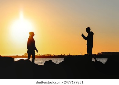 Silhouette of back lit young woman posing on harbor rock wall for photograph taken by young man with mobile phone at sunset. Two tone, orange and black. - Powered by Shutterstock