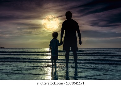 Silhouette back view of loving child holding hands her father, standing in the sea. Family enjoying and relaxing on beach with full moon on sky background. The moon taken with my own camera.