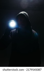 Silhouette of bandit in hoodie holding flashlight on black background with smoke