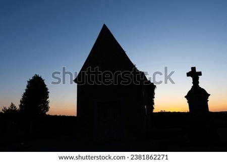 silhouette of basic rustic church in graveyard at sunset. tree and gravestone with crucifix cross either side of small worship cemetery building. Bold tombstone in front of dusk sky