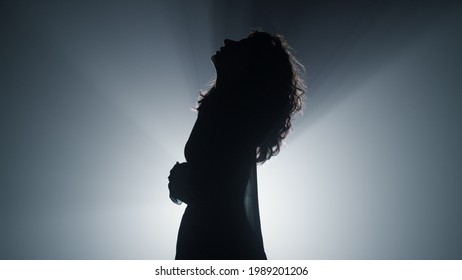 Silhouette of beautiful girl standing in spotlight background. Side view of young woman looking up in dark space. Attractive female person moving hands in air indoors.  - Powered by Shutterstock