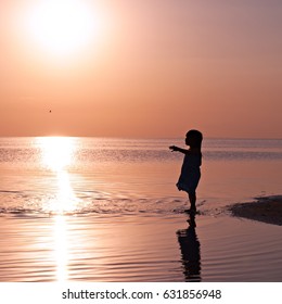 Silhouette Of A Beautiful Little Girl Standing In Water On Sunset Beach. Very Beautiful Sunset At The Seaside