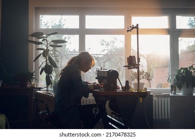 Silhouette of a beautiful middle aged tailor woman sewing on a sewing machine while sitting at her moody working place at home. Fashion atelier, tailoring, handmade clothes concept. Slow Fashion. - Powered by Shutterstock