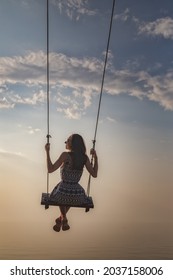 Silhouette of a beautiful young girl who is swinging on a swing against the background of the sky and sea waves. Real photo without graphic editing.