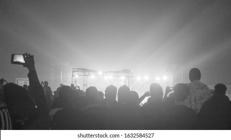 Silhouette Of Big Audience Crowd Of Happy People Standing At Concert Grounds Cheering Clapping Use Smart Phones Raising Hands Up In Air Facing Music Band Enjoying. Stage Light Reflection In Front.