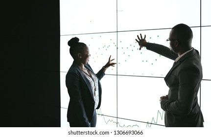 Silhouette Of A Black African Business People Giving A Presentation Conference. Pointing At A Large Video Screen With Charts And Graphs Next To Her In A Dark Room