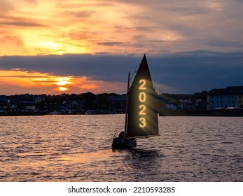Silhouette of a boat sailing into town. Glowing sign 2023 on the sail of the yacht. Celebrating new year concept. Dramatic cloudy sky in the background with rising sun. - Shutterstock ID 2210593285